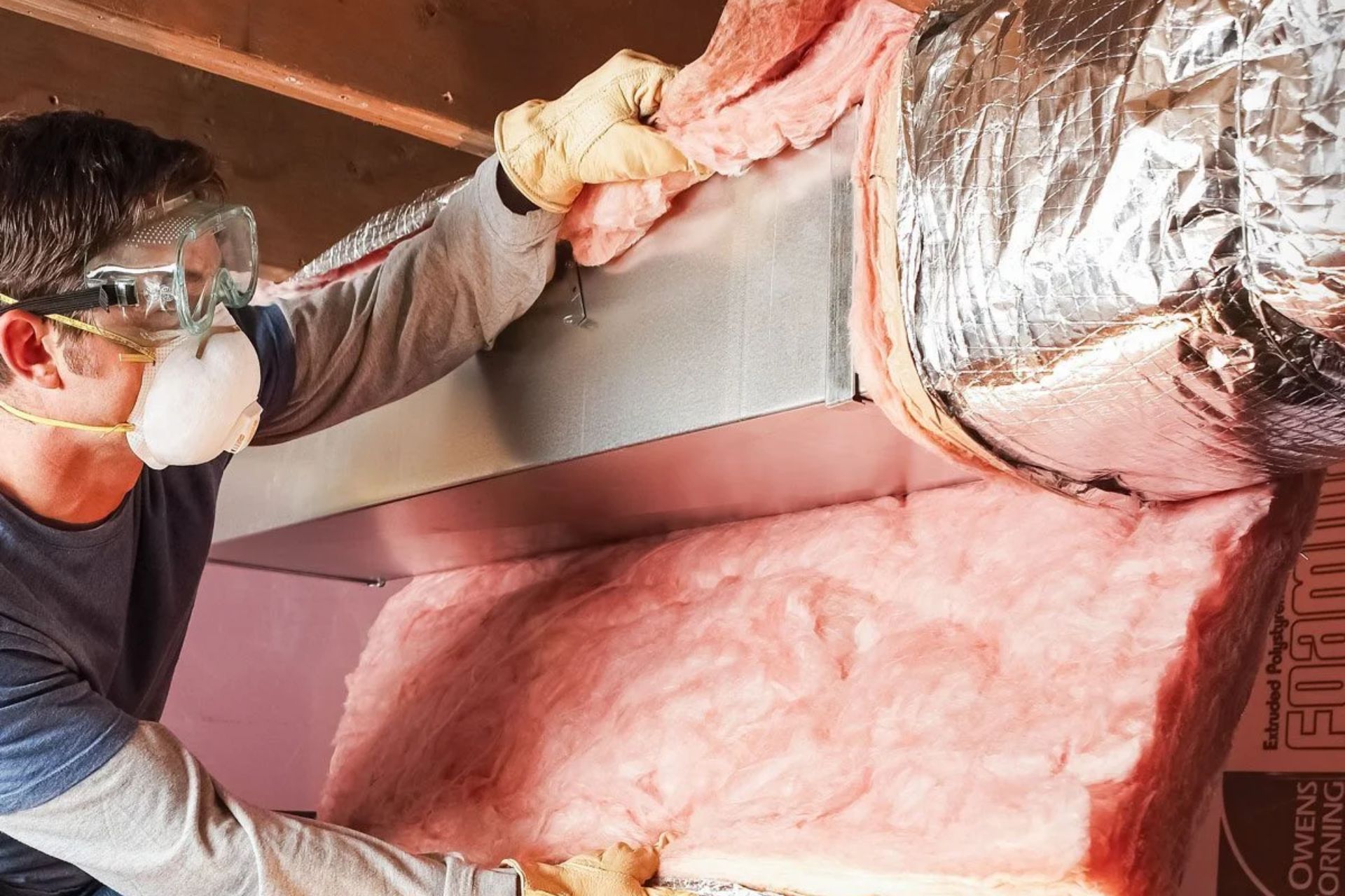 A man wearing a mask and gloves is seen installing insulation, highlighting safety measures in a construction environment.