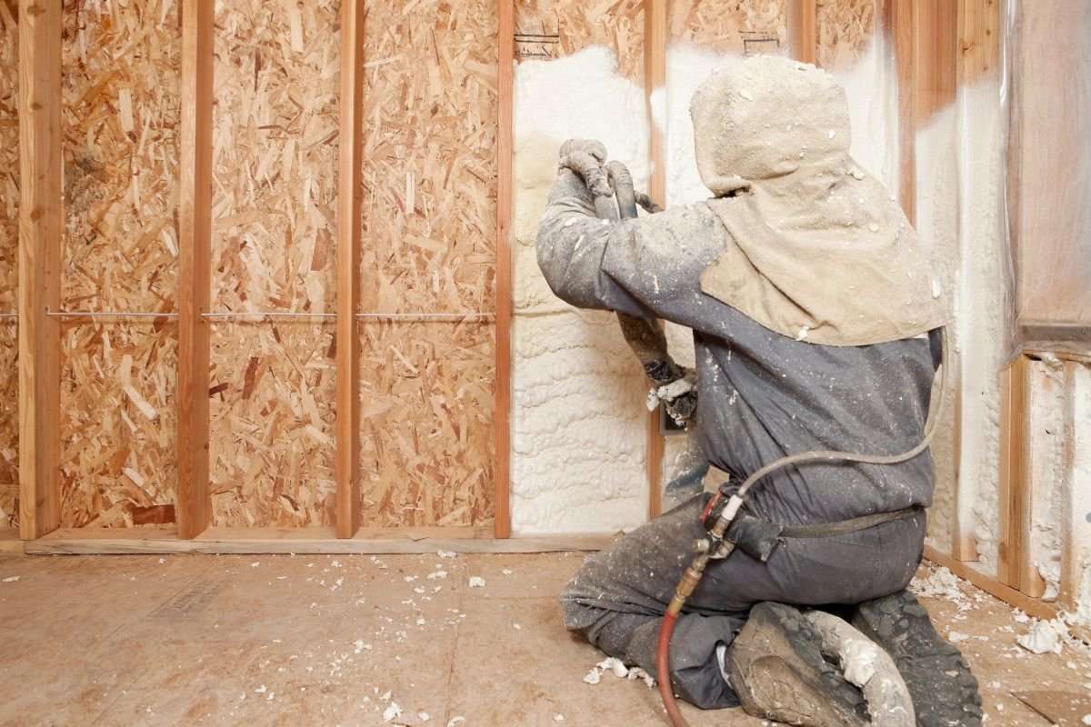 A worker wearing a hard hat and jacket applies paint to a wall during an insulation service task.